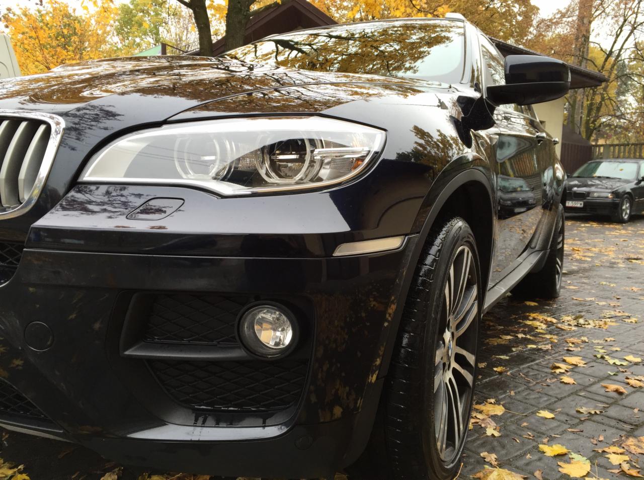 Ciemny SUV, widok z przodu, po umyciu, z widocznymi kroplami wody na karoserii, na tle jesiennych liści na brukowanej nawierzchni.