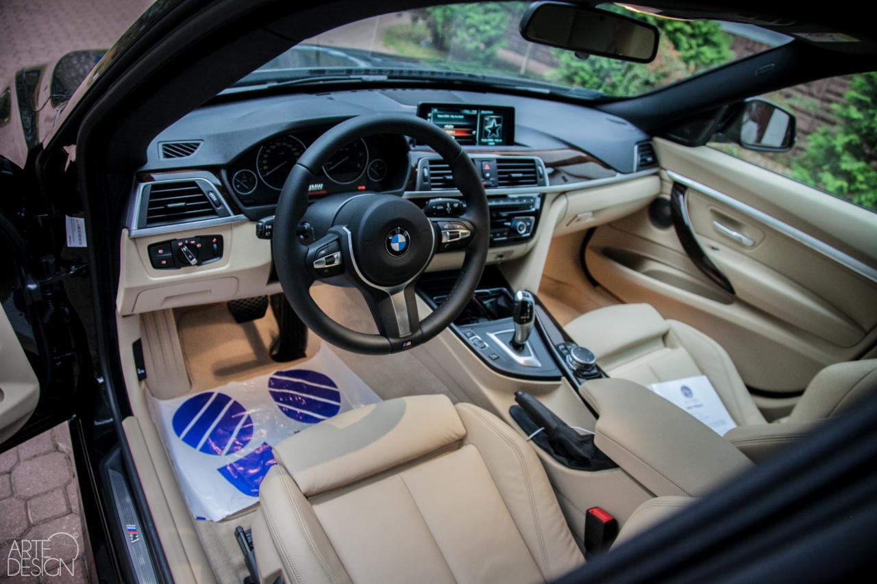 Wnętrze czarnego BMW serii 4 Cabrio z jasną skórzaną tapicerką, widoczne z zewnątrz przez otwarte drzwi, z ochronną folią na dywanikach.