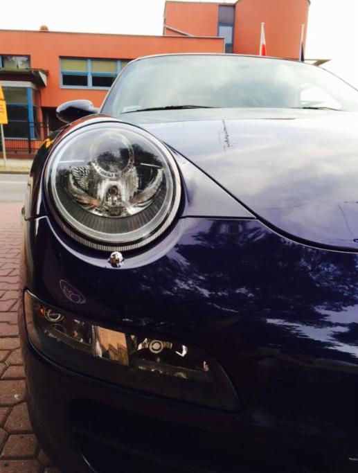 Zbliżenie na reflektor i fragment karoserii granatowego Porsche, widoczne odbicie otoczenia w lakierze.