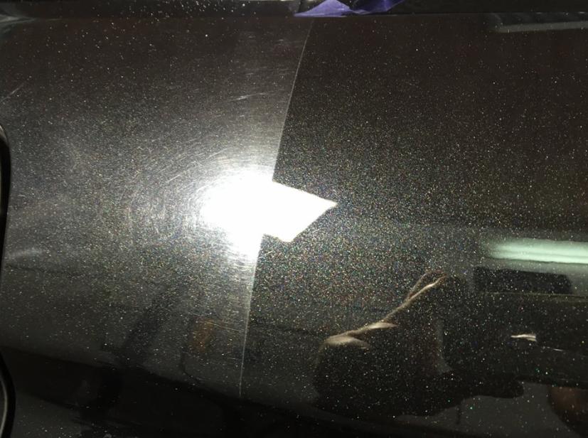 Część karoserii samochodu w kolorze ciemnego grafitu, po lewej stronie widoczne zarysowania i niedoskonałości lakieru, po prawej stronie fragment po polerowaniu lakieru, z wyraźnym odbiciem światła.