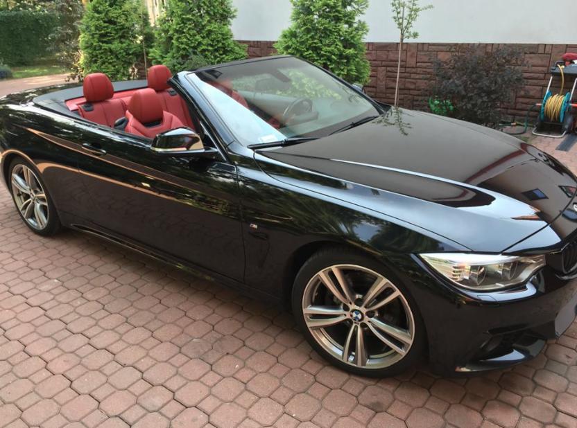 Błyszczący czarny kabriolet BMW z czerwonym skórzanym wnętrzem na tle brukowanej posesji w Otwocku.