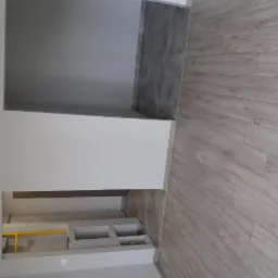 Wykończone wnętrze: jasne panele podłogowe imitujące drewno, szare płytki na ścianie, białe ściany z wnękami, widoczna instalacja z żółtą rurką.