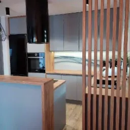 Nowoczesna kuchnia w odcieniach szarości z drewnianymi akcentami, wysoka czarna tuba okapu, drewniana ażurowa ścianka działowa i drewniany blat wyspy kuchennej.