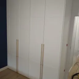 Biała szafa wnękowa z czterema drzwiami i złotymi uchwytami, na podłodze panele w odcieniach brązu, ściana w kolorze ciemnego granatu, widok z boku.