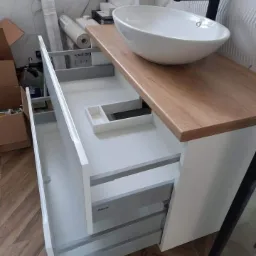 Biała szafka łazienkowa z drewnianym blatem, owalną umywalką nablatową i otwartymi szufladami z systemem Blum, widoczne materiały budowlane w tle.