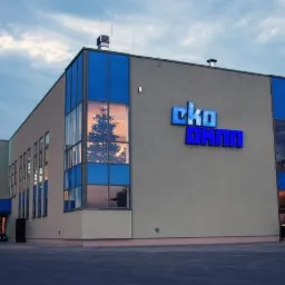 Budynek firmy Eko Okna z niebieskimi ramami okiennymi i logo firmy, w oknach widoczne odbicie drzewa.