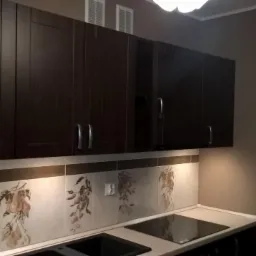 Ciemnobrązowe szafki kuchenne podwieszane z metalowymi uchwytami, ceramiczna mozaika z motywem owocowym nad blatem, płyta indukcyjna.