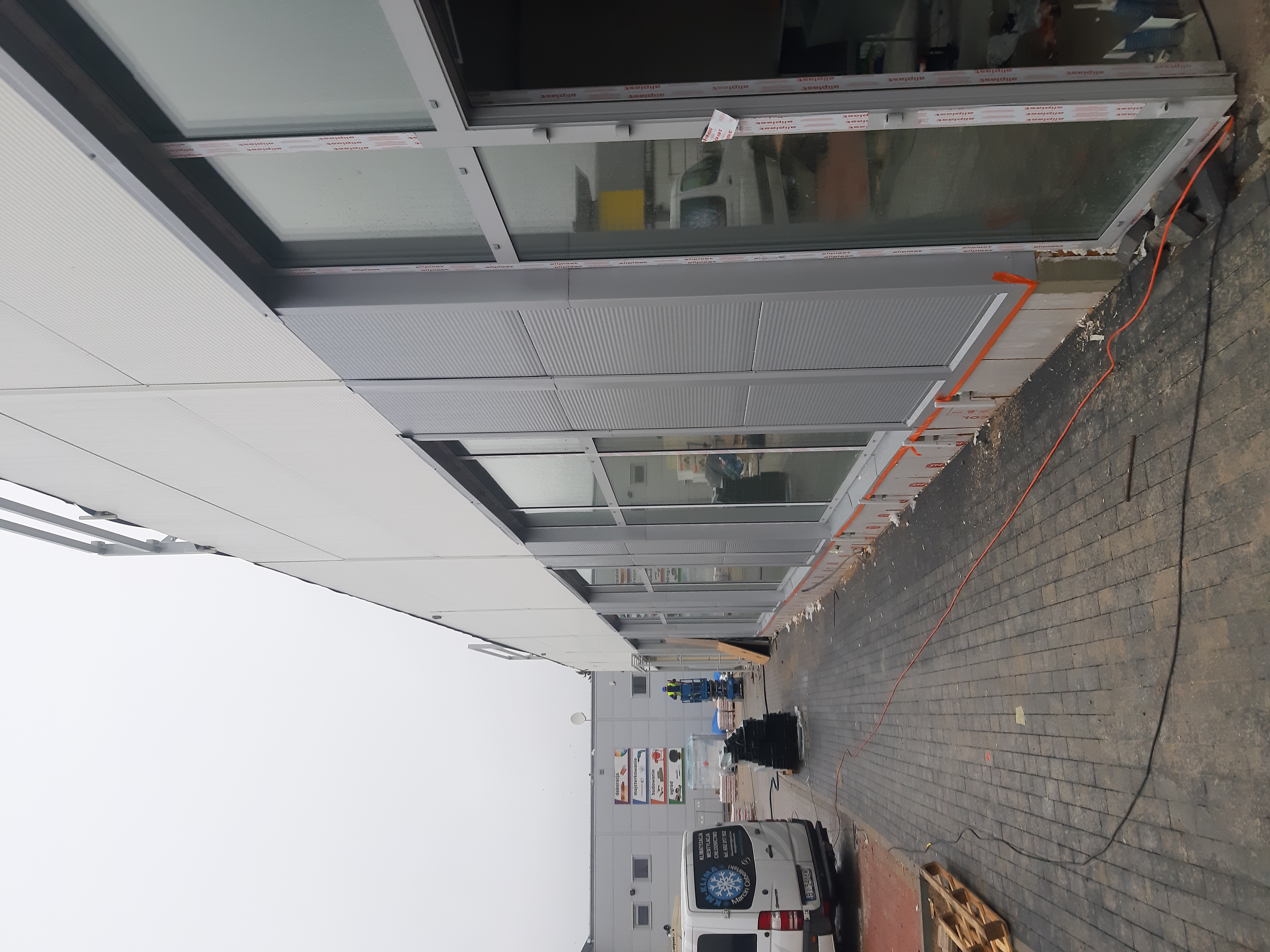 Budynek hali w trakcie montażu okien i obudowy z płyt warstwowych, widoczny podnośnik koszowy z pracownikami, materiały budowlane i samochód dostawczy firmy klimatyzacyjnej.