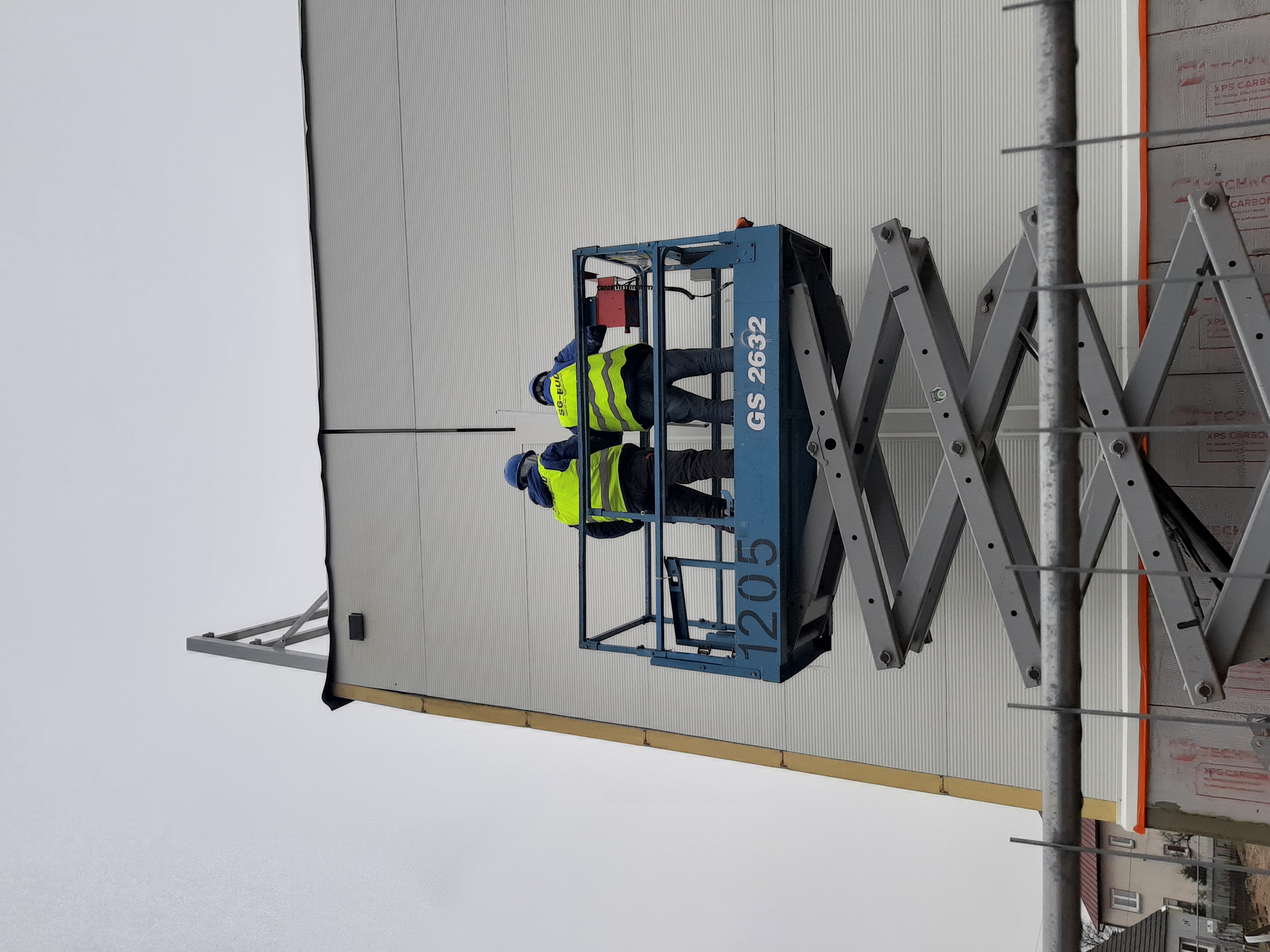 Dwóch pracowników w żółtych kamizelkach i niebieskich kaskach na podnośniku nożycowym, montujących jasnoszare płyty warstwowe na ścianie hali. Widoczne detale konstrukcji stalowej i sąsiednie budynki.