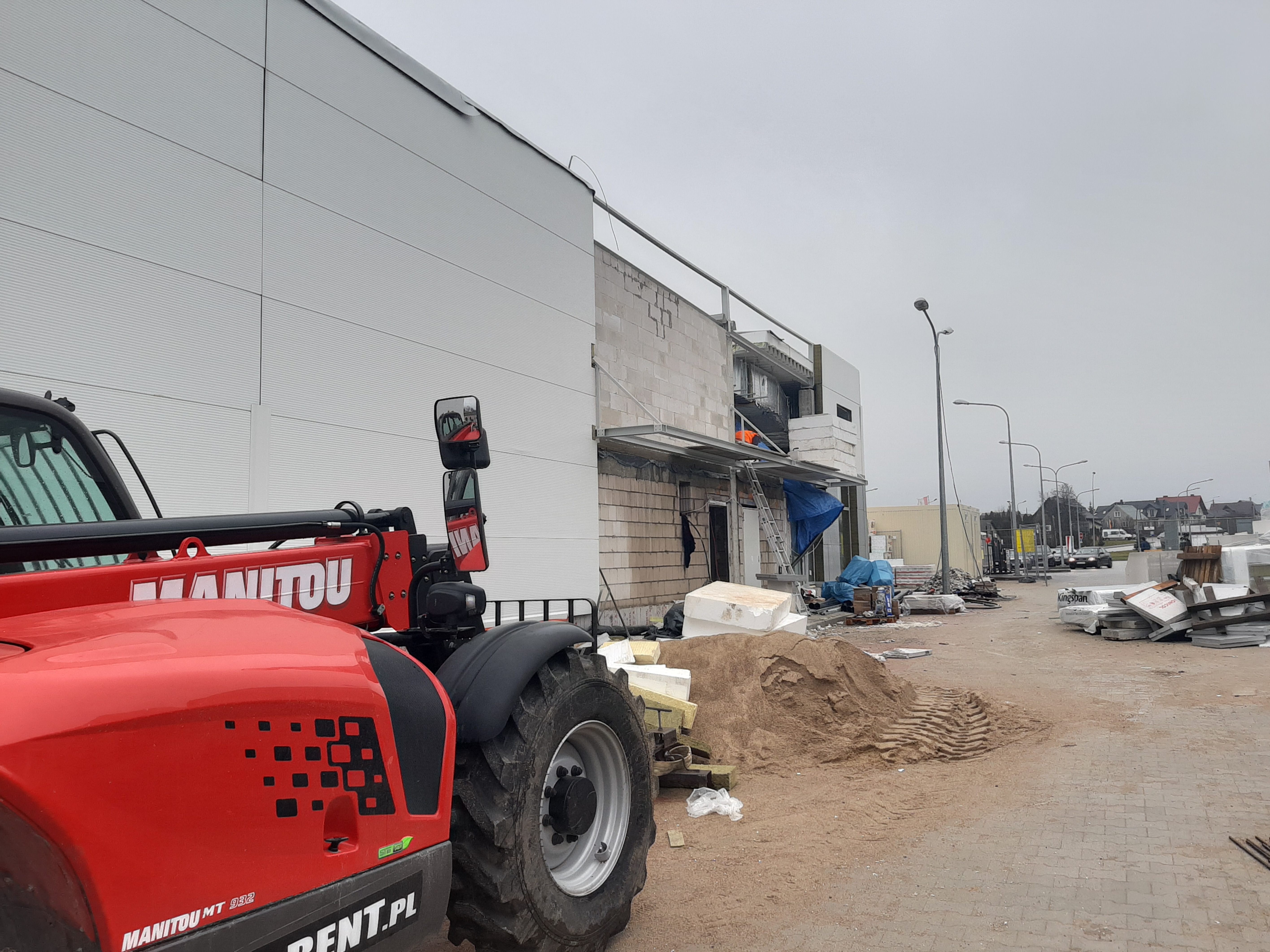 Czerwony podnośnik teleskopowy Manitou MT 932 na placu budowy hali z białych płyt warstwowych, widoczne materiały budowlane i pracownik w pomarańczowej kamizelce w wejściu.