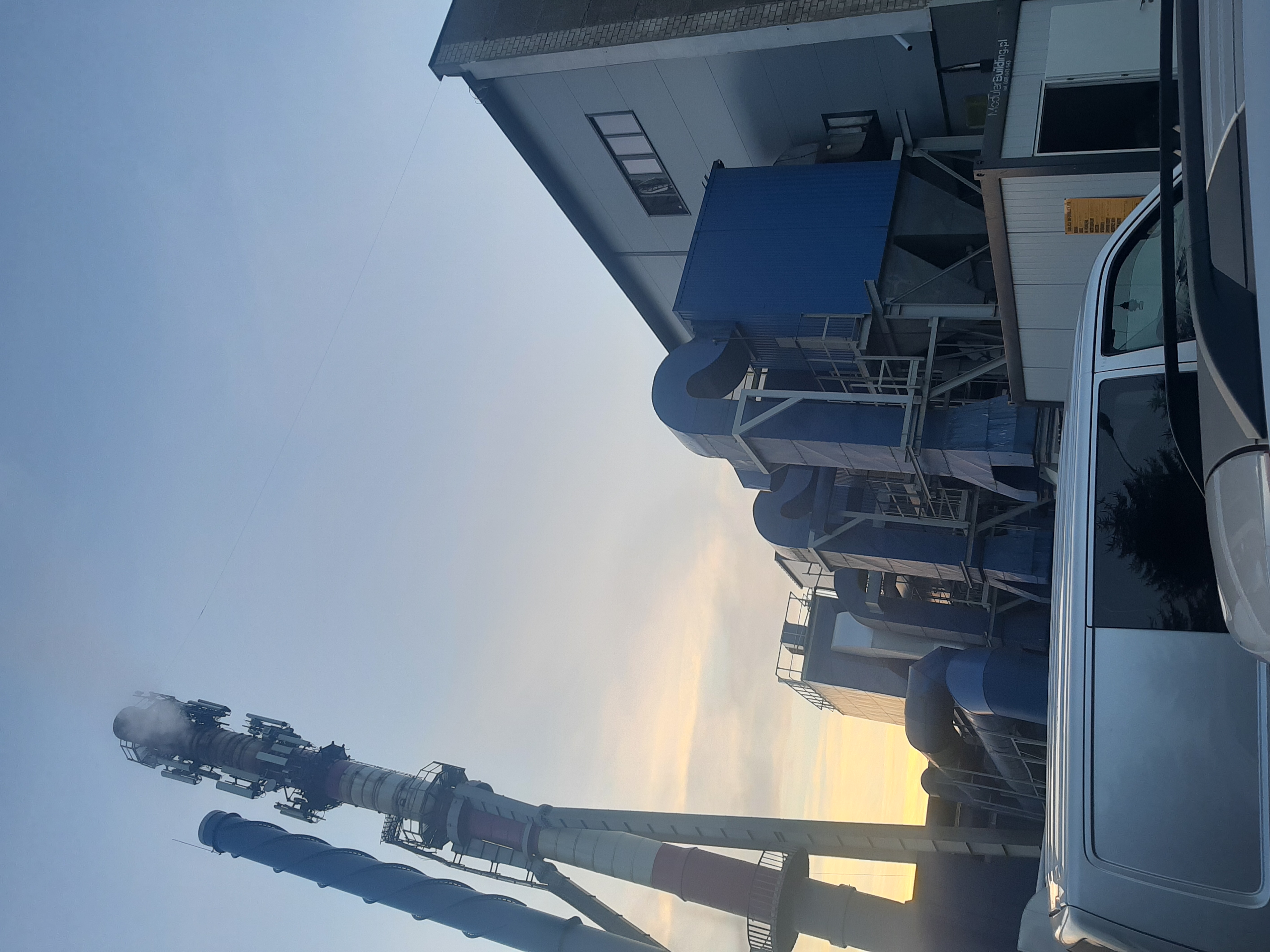 Widok na przemysłową halę z niebieskimi elementami wentylacyjnymi i kominem z instalacją telekomunikacyjną, sfotografowaną z perspektywy pojazdu.