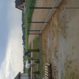 Widok na nowo postawione ogrodzenie panelowe z metalową siatką na betonowej podmur&oacute;wce w pochmurny dzień, z kałużami na ziemi i widocznym budynkiem w tle.