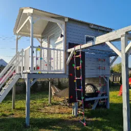 Drewniany plac zabaw w Dębie z domkiem na podeście, schodami, ścianką wspinaczkową i drabinką sznurkową, ozdobiony kolorowymi lampionami, na tle zielonego trawnika.