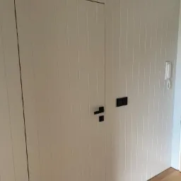 Ukryte, białe drzwi z pionowymi panelami, z czarną klamką i włącznikiem światła, wkomponowane w ścianę z panelami, na drewnianej podłodze.