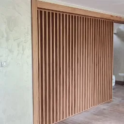 Drewniana ściana działowa z pionowych listew w jasnym odcieniu drewna, tworząca przesuwne drzwi w nowoczesnym wnętrzu, widoczna błękitna szklana tafla drzwi z motywem drzewa w tle.