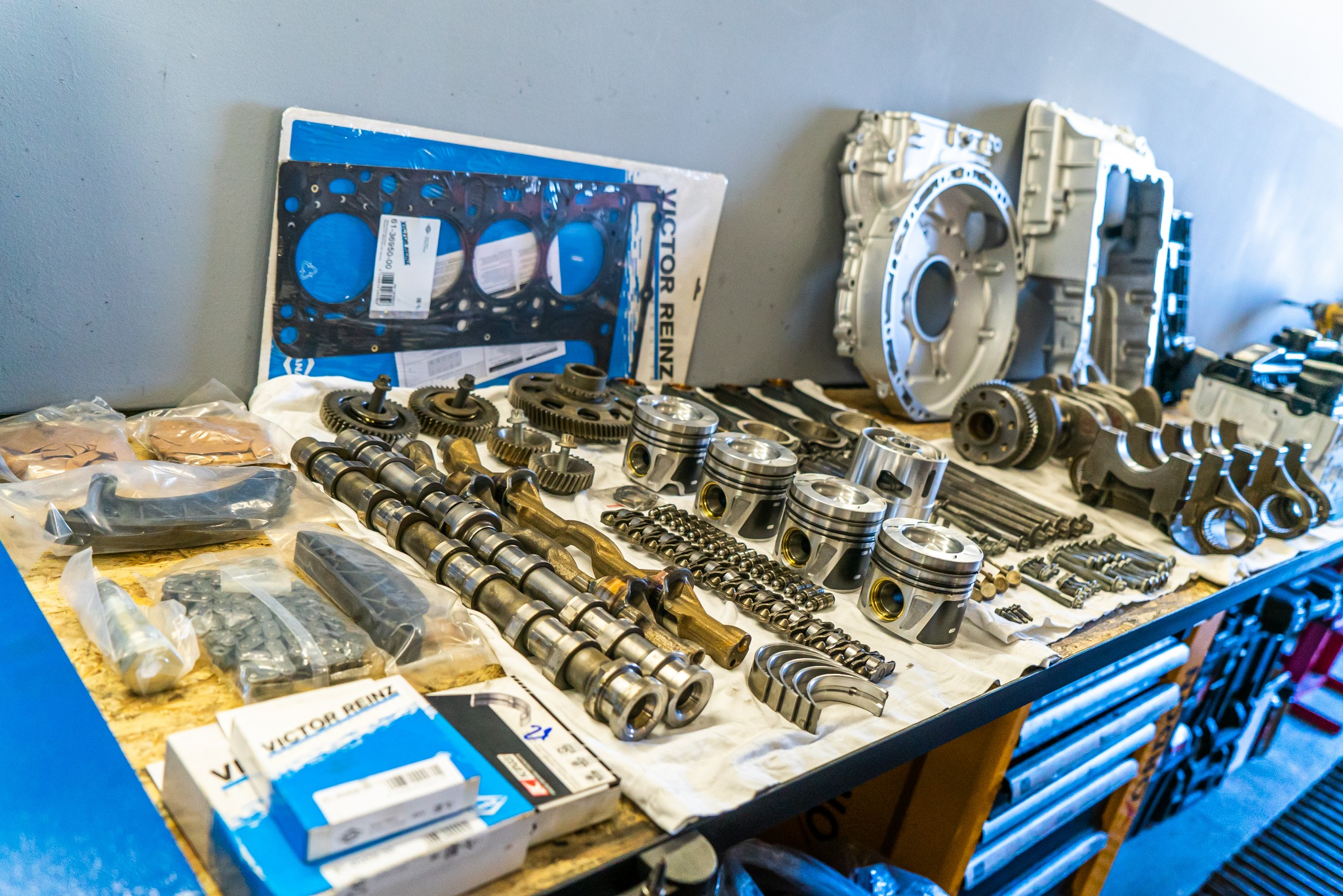 Różne części samochodowe, w tym tłoki, wałki rozrządu, przekładnia i uszczelki, ułożone na stole warsztatowym przed montażem.