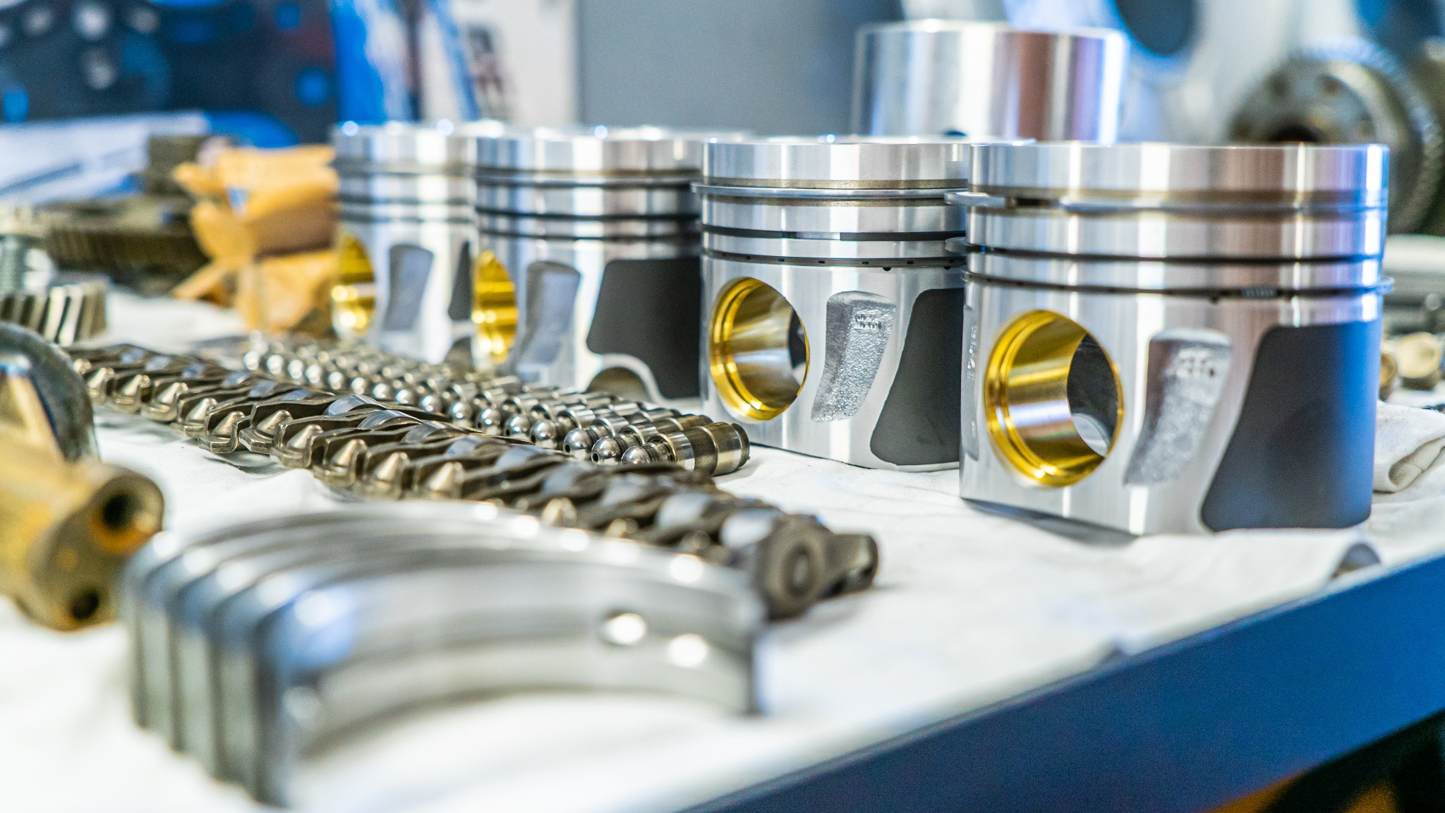 Rząd czterech srebrnych tłoków silnika z wyraźnymi złotymi otworami, wałek rozrządu i inne części zamienne na białym stole w warsztacie w Czarnej Sędziszowskiej.