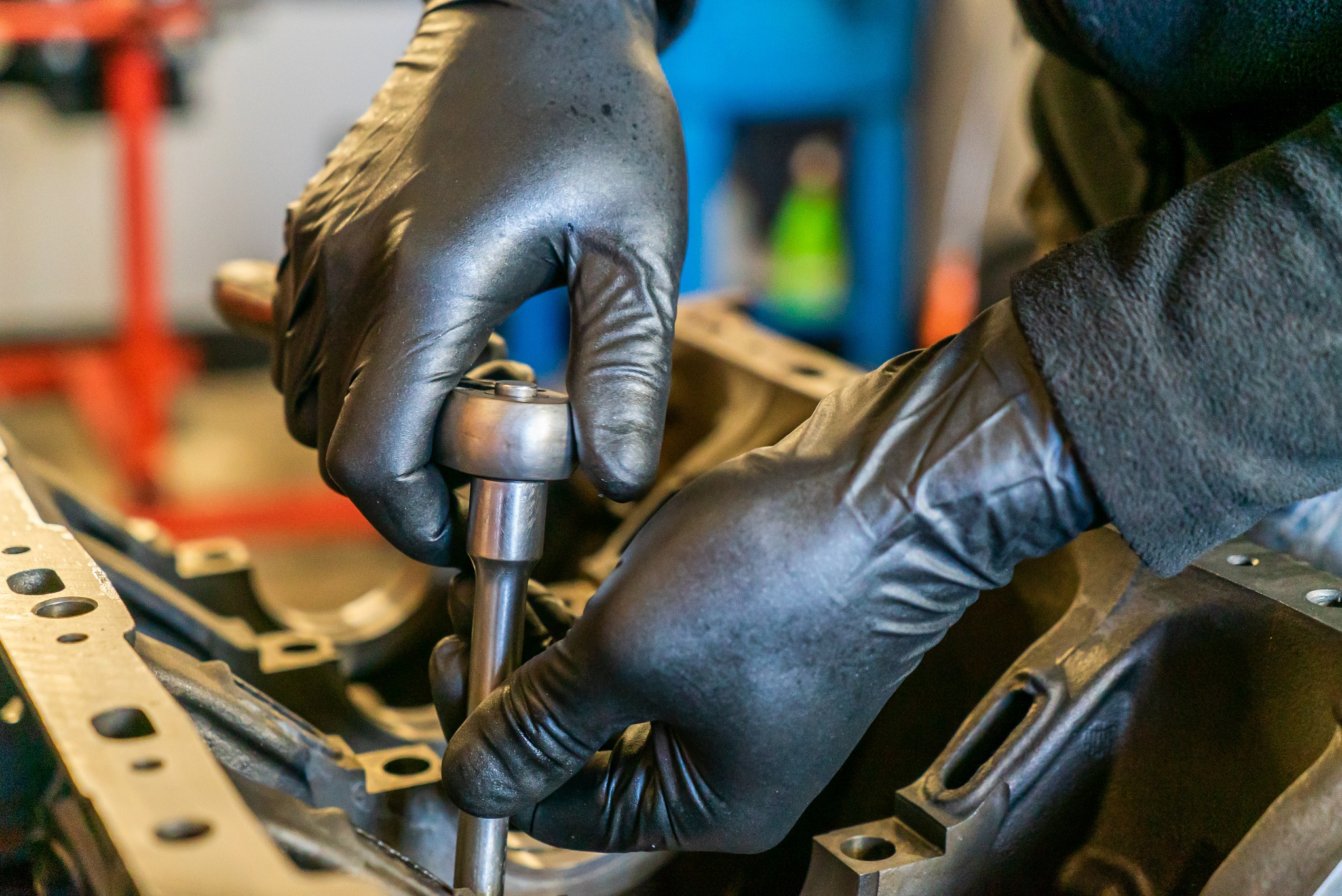Ręce w czarnych rękawicach skręcają element silnika samochodowego za pomocą klucza nasadowego, widoczny fragment bloku silnika.