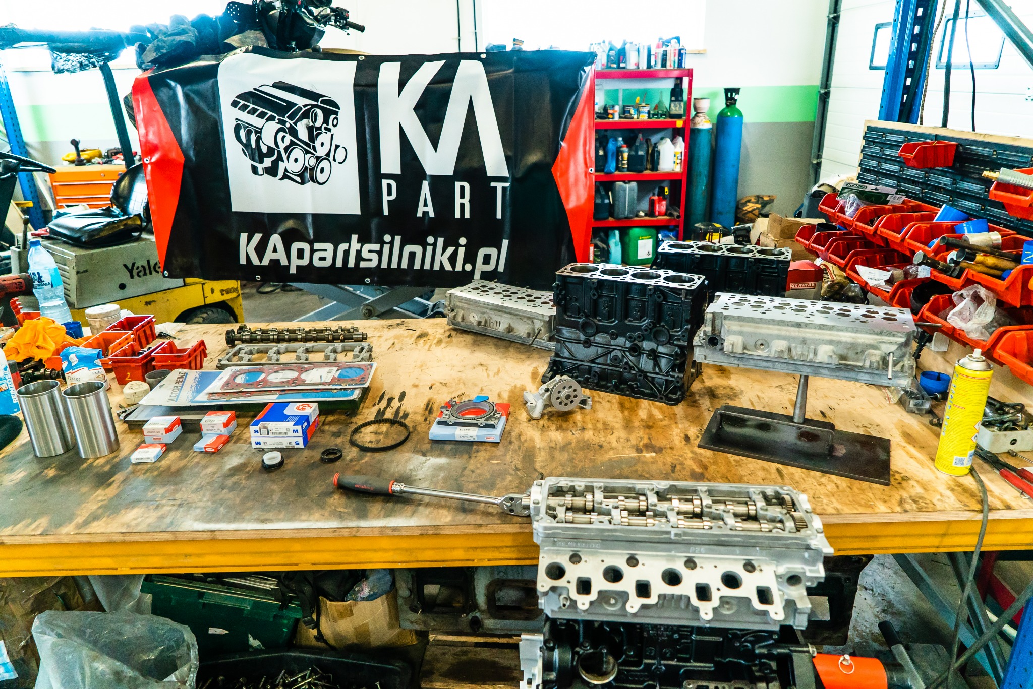 Warsztatowy stół roboczy z rozłożonymi częściami silnika samochodowego, głowice cylindrów, blok silnika, uszczelki, narzędzia, w tle baner reklamowy z logo KA Parts i regał z olejami.
