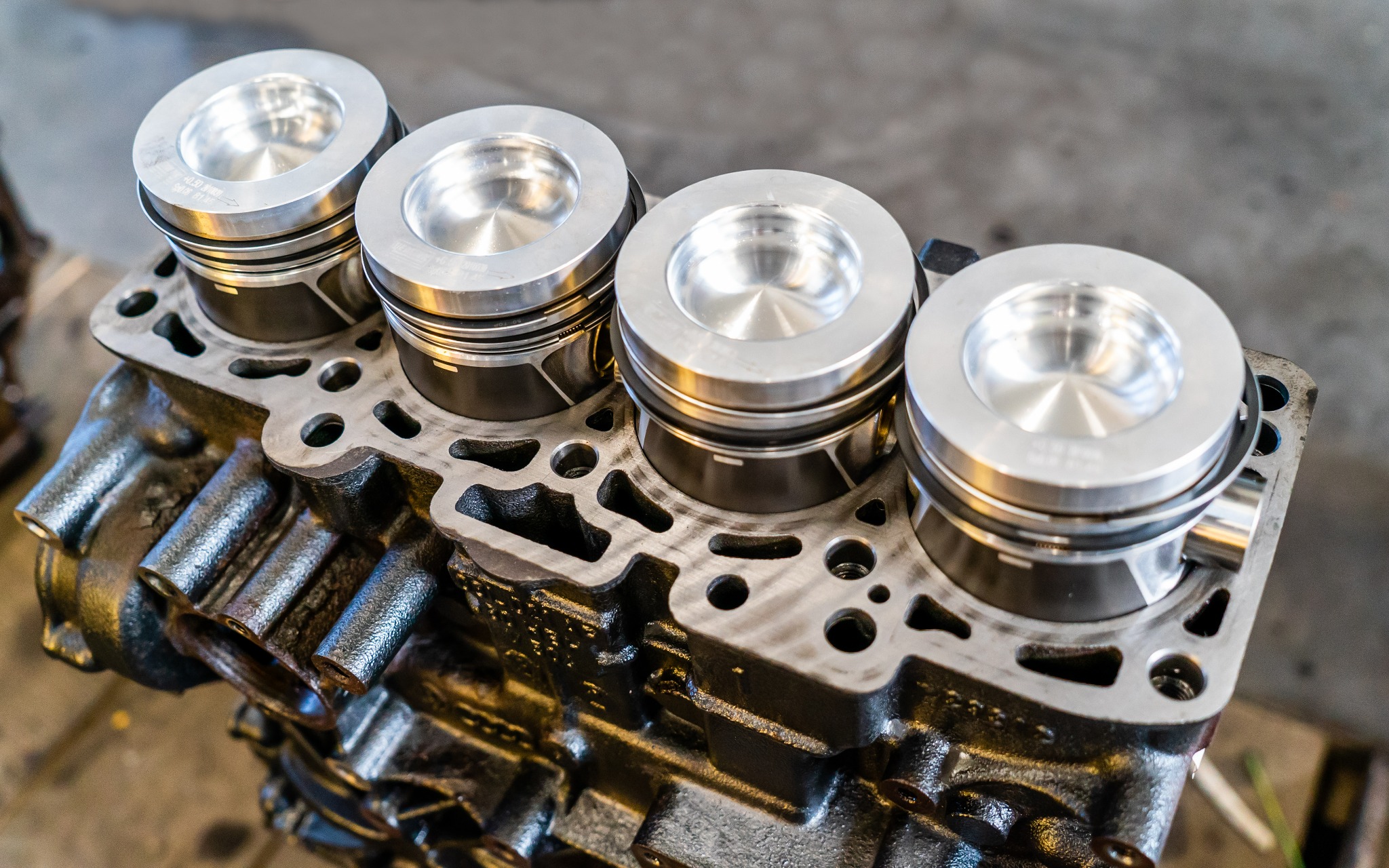 Blok silnika z czterema nowymi, srebrnymi tłokami zamontowanymi, widoczne detale konstrukcyjne i oznaczenia na tłokach.