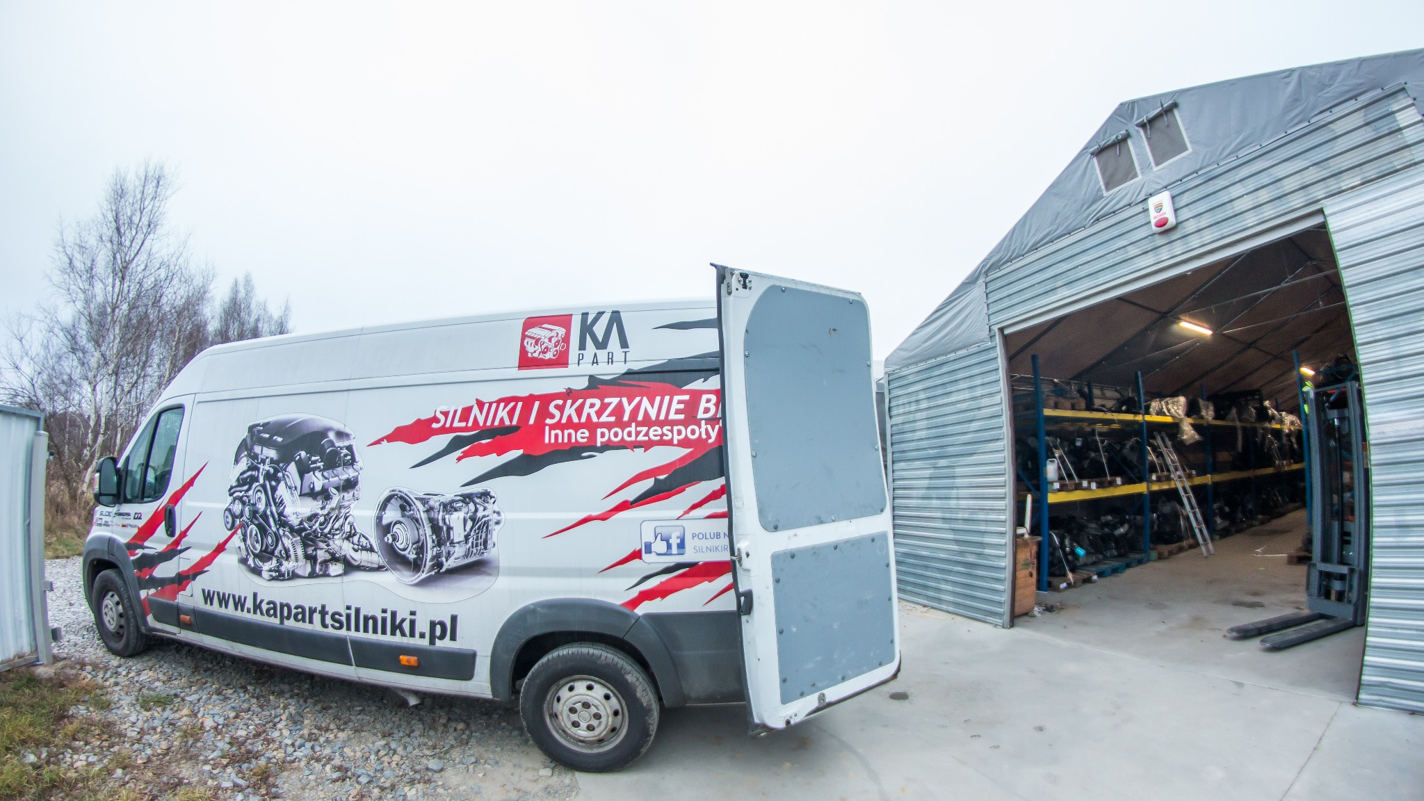 Furgonetka z grafiką silnika i skrzyni biegów parkuje przed magazynem z regałami pełnymi części samochodowych.
