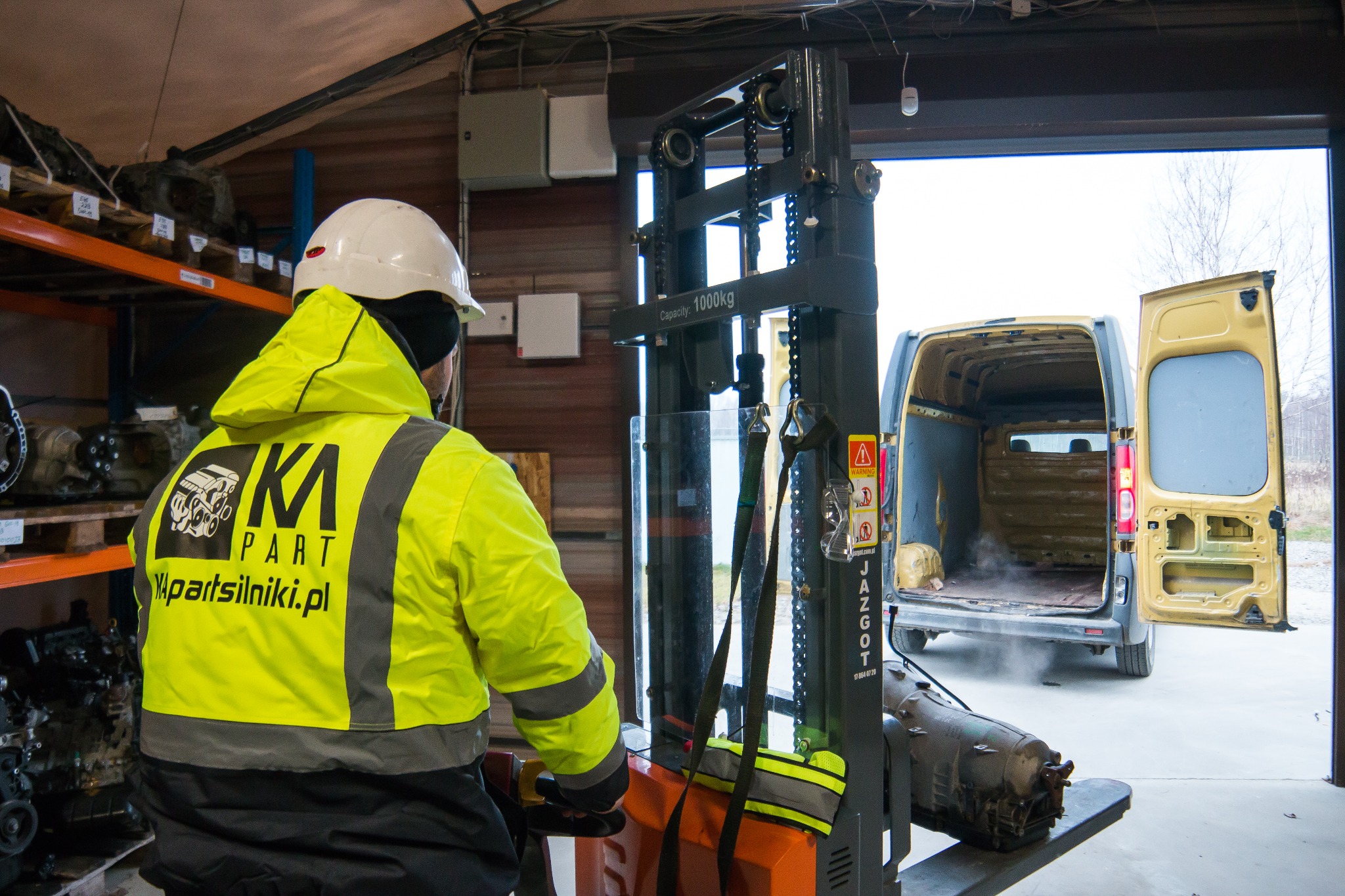 Pracownik w żółtej kurtce z logo firmy transportuje wózkiem widłowym skrzynię biegów w warsztacie samochodowym, w tle otwarty furgon.