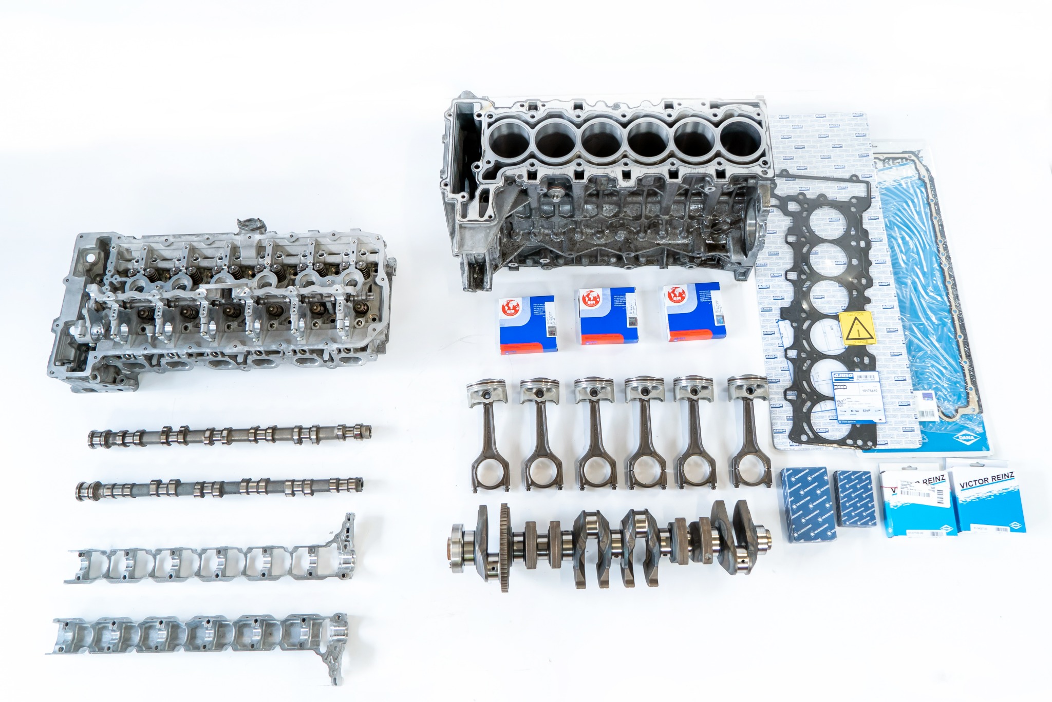 Rozłożony silnik samochodowy na białym tle, widoczne: blok silnika, głowica, wałki rozrządu, tłoki z korbowodami, wał korbowy, uszczelki i opakowania filtrów, przygotowane do montażu.