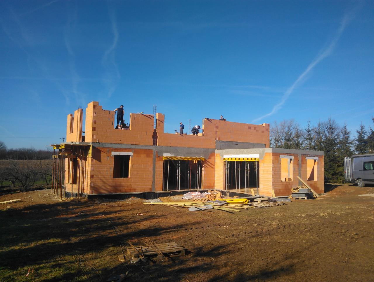 Wznoszenie ścian domu murowanego z pustaków ceramicznych; widoczne elementy szalunków okiennych i stropu oraz robotnicy na dachu, na tle błękitnego nieba i drzew