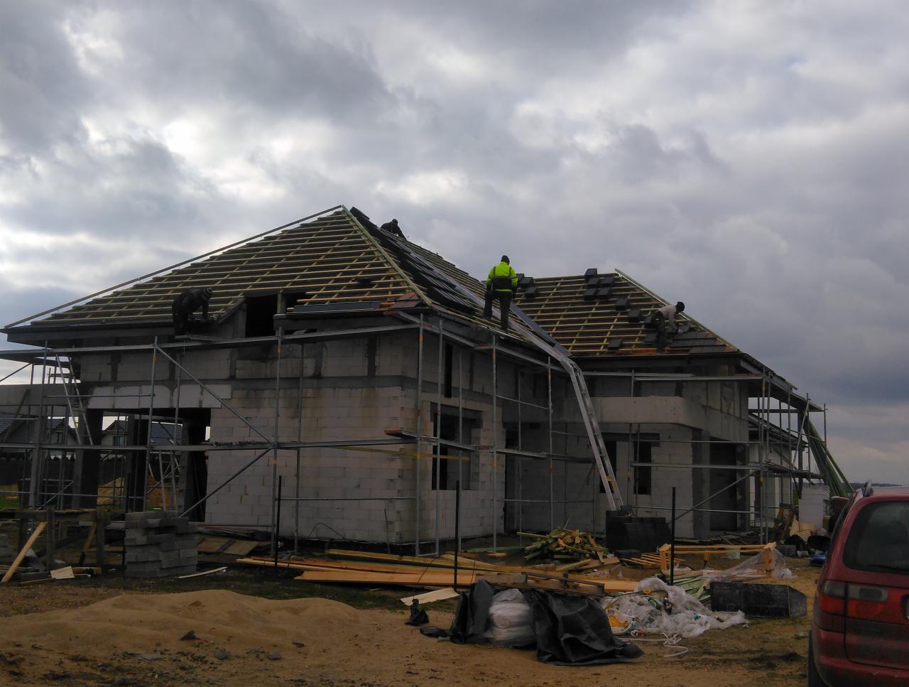 Budowa domu jednorodzinnego z białego pustaka: montaż drewnianej więźby dachowej i układanie czarnej dachówki przez robotników w jaskrawych kamizelkach na tle pochmurnego nieba.