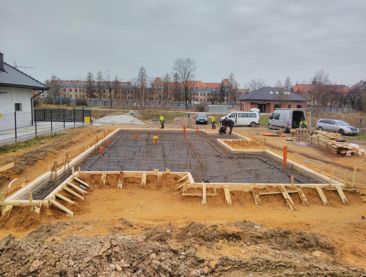 Zbrojenie fundamentu domu jednorodzinnego z szalunkiem drewnianym i wystającymi rurami wentylacyjnymi, widoczne samochody i budynki w tle.