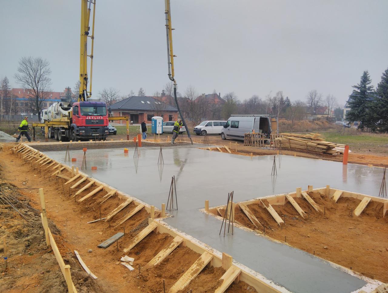 Wylewanie betonu na przygotowany szalunek fundamentów domu jednorodzinnego przy użyciu pompy do betonu Jetlift Man, widoczni pracownicy w odblaskowych kamizelkach, w tle budynki mieszkalne...