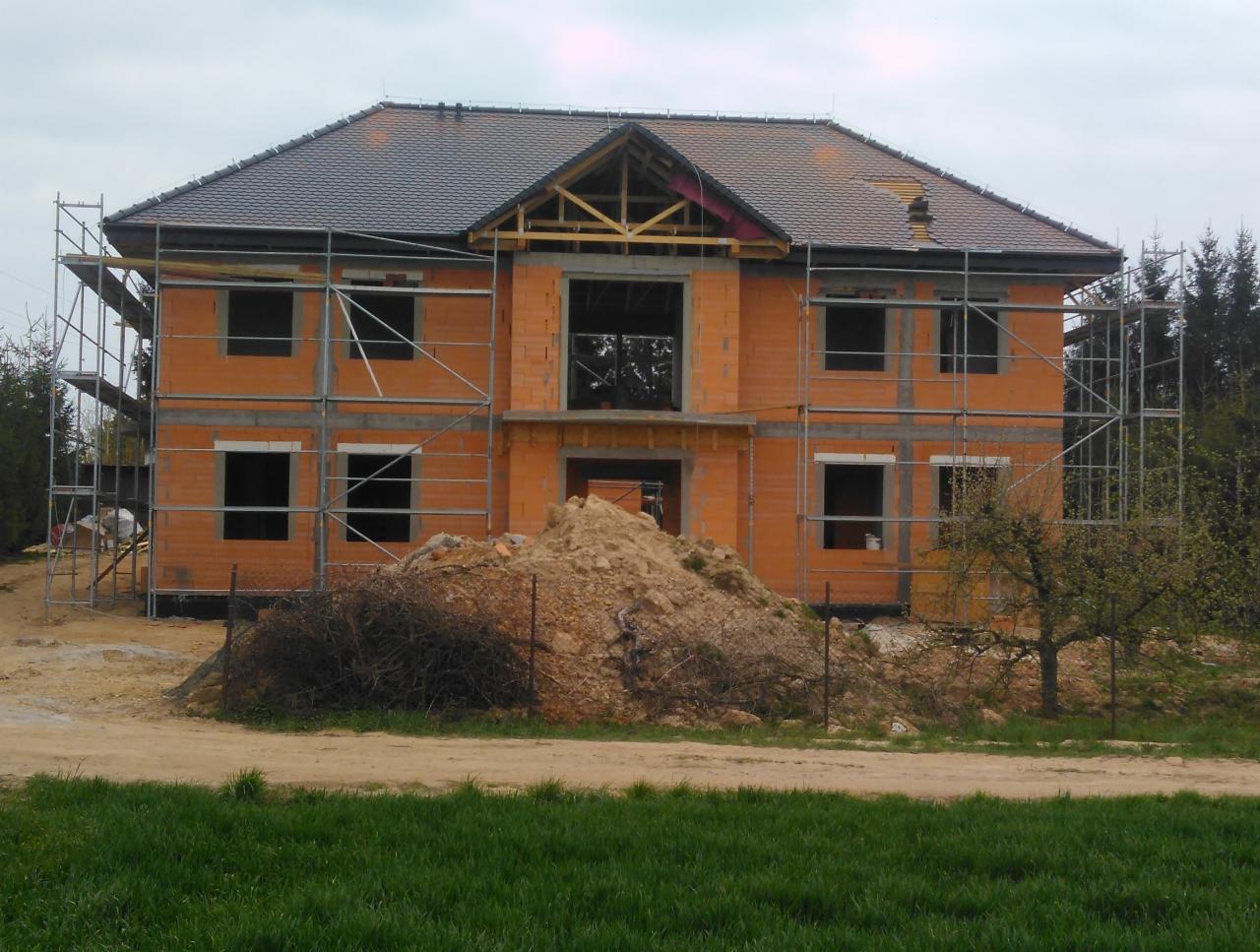 Dwukondygnacyjny budynek w trakcie wznoszenia z czerwonej cegły, otoczony rusztowaniami, z zamontowaną więźbą dachową i pokryciem z ciemnej dachówki, na pierwszym planie hałda ziemi i trawa.