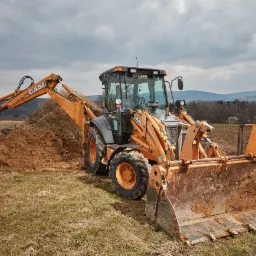 Pomarańczowa koparko-ładowarka CASE podczas prac ziemnych na polu, z widocznym stosem ziemi i odległym krajobrazem górskim pod pochmurnym niebem.