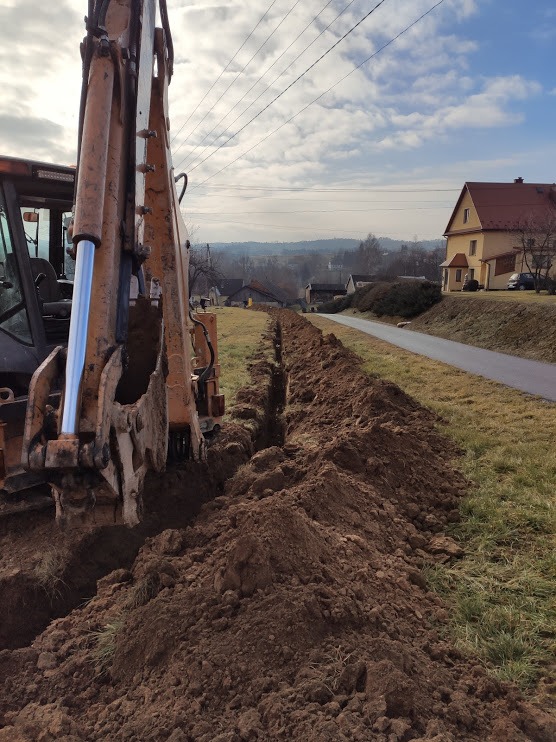 Koparka wykopuje wąski rów w ziemi wzdłuż drogi asfaltowej, widok na pagórkowaty krajobraz z zabudowaniami w tle pod błękitnym niebem.