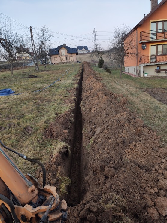 Wąski wykop liniowy w ziemi, wykonany prawdopodobnie pod instalację, z widocznym fragmentem koparki i budynkami mieszkalnymi w tle.
