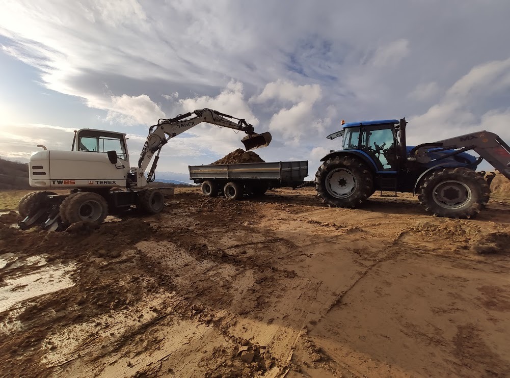 Biała koparka Terex ładuje ziemię na przyczepę traktora New Holland w trakcie prac ziemnych na placu budowy przy pochmurnym niebie.