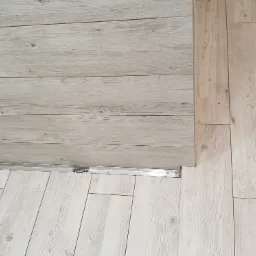 Ułożona pod kątem podłoga z płytek imitujących drewno w odcieniach bieli i beżu, z widocznym odpływem liniowym i fragmentem ściany wyłożonej płytkami imitującymi biały marmur.