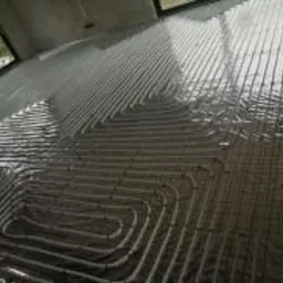 Równomiernie rozłożone rury grzewcze na folii izolacyjnej, przygotowane do zalania betonem w surowym wnętrzu z widocznym oknem.