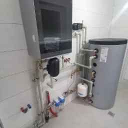 Nowoczesna instalacja grzewcza z zasobnikiem i filtrami wody, widok na białe płytki i szare elementy systemu.