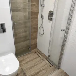 Nowoczesna łazienka z kabiną prysznicową typu walk-in, podłogą imitującą drewno i białą armaturą, widoczny odpływ liniowy.