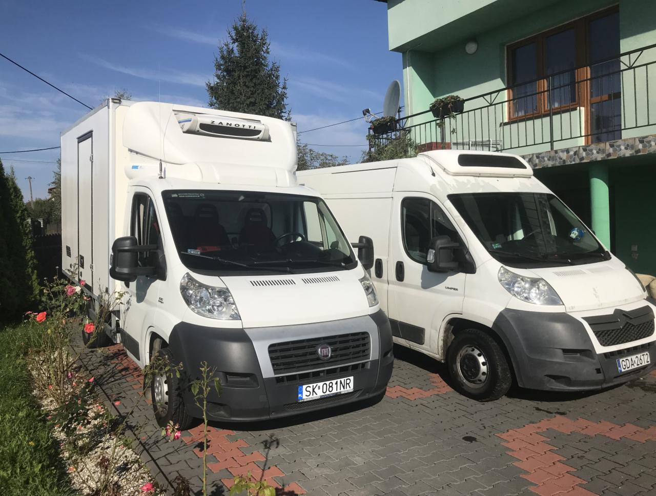 Dwa białe busy dostawcze zaparkowane na brukowanym podjeździe przed zielonym domem, jeden z chłodnią Zanotti.