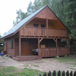 DOM-LET Usługi Ciesielskie Roman Ryżak - Dwukondygnacyjny drewniany dom z balkonem, widok z zewnątrz, zielony dach, otoczenie drzewa, tablica informacyjna z prawej strony.