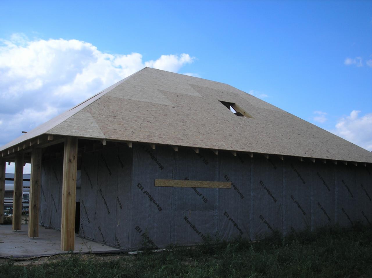 Budowa domu: widok na konstrukcję ścian z membraną izolacyjną NovoRoof 115 i fragment dachu z otworem, drewniane słupy podtrzymujące zadaszenie.