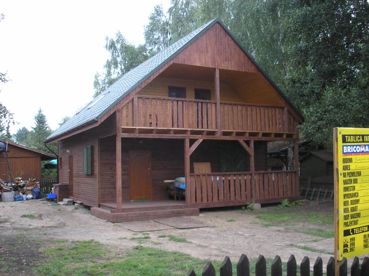 Dwukondygnacyjny drewniany dom z balkonem, widok z zewnątrz, zielony dach, otoczenie drzewa, tablica informacyjna z prawej strony.