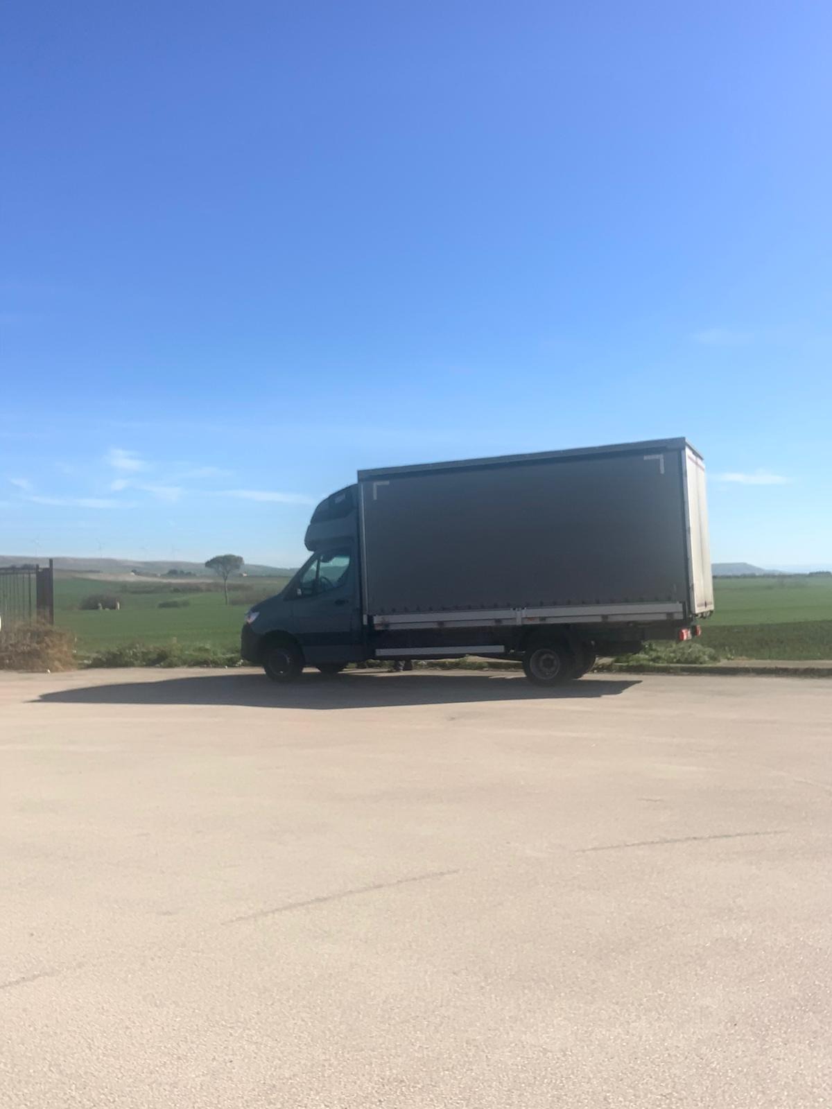 Szary bus dostawczy z plandeką zaparkowany na betonowym placu, w tle zielone pola i błękitne niebo.