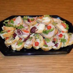 Czarny, ośmiokątny p&oacute;łmisek wypełniony r&oacute;żnorodnymi, apetycznymi kanapeczkami na jasnym pieczywie z sezamem, udekorowanymi świeżymi warzywami, wędlinami i kiełkami, ułożony na drewnianym blacie.