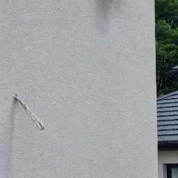 Kamera monitoringu przymocowana do jasnej, teksturowanej ściany budynku, widoczny fragment dachu sąsiedniego budynku i zieleń drzew w tle, obok wystający element mocujący.