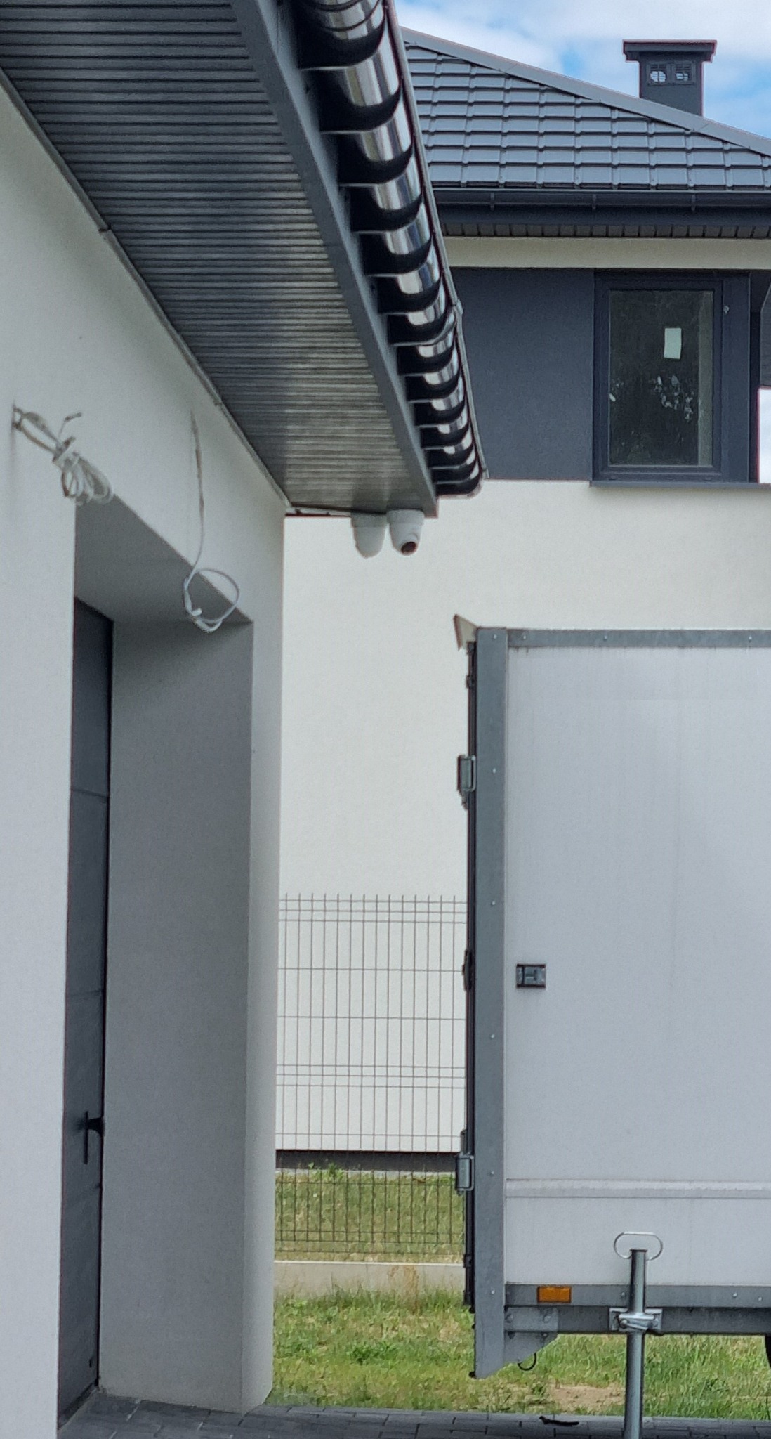 Dwie białe kamery monitoringu zamontowane pod okapem domu, z widocznymi kablami i fragmentem zaparkowanej przyczepy transportowej.