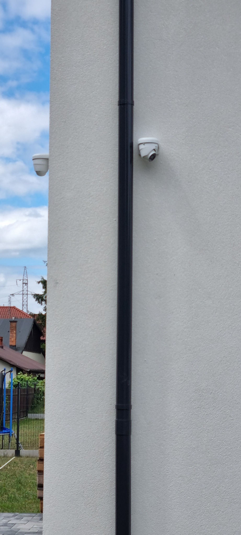 Dwie białe kamery kopułkowe zamontowane na elewacji budynku, obok pionowej rury spustowej, z widocznym fragmentem sąsiedniego domu i niebem w tle.