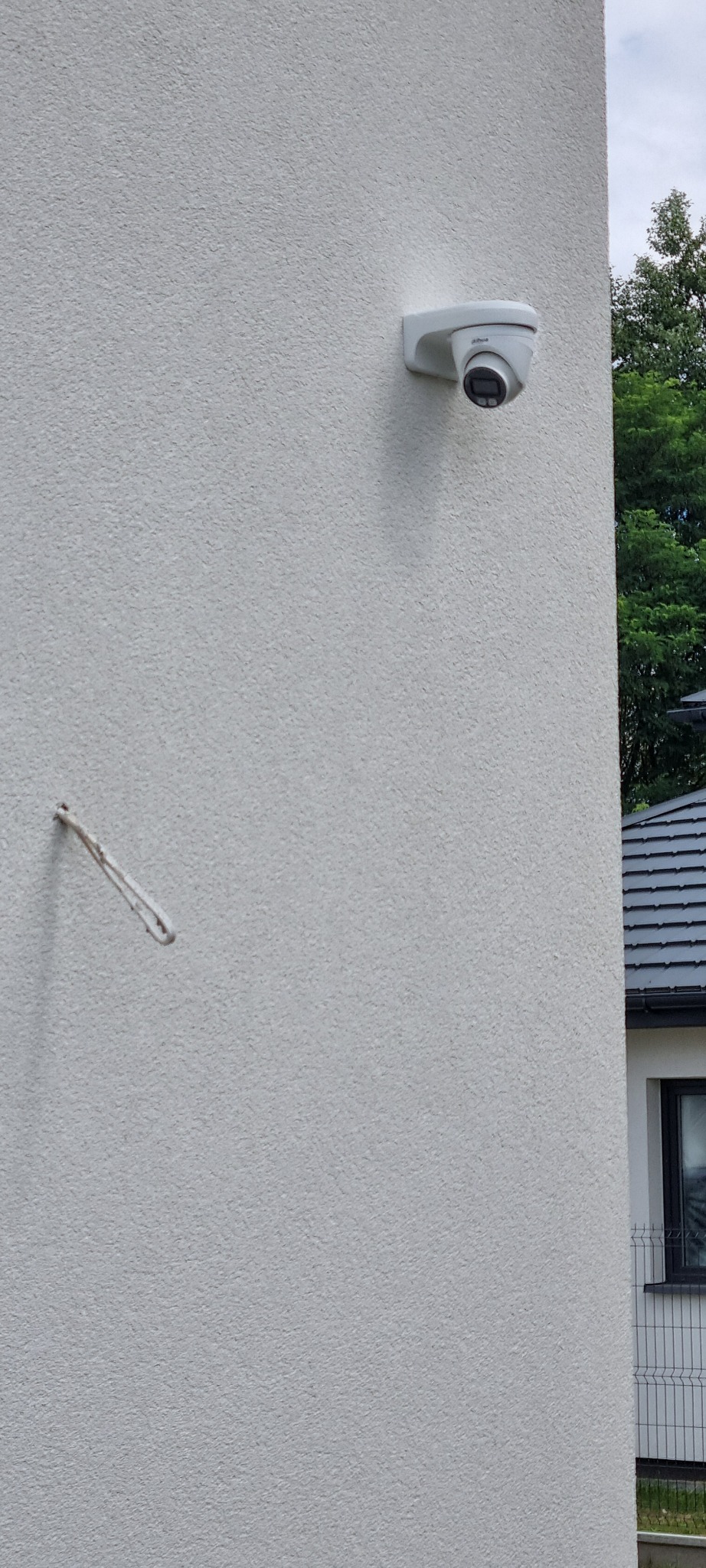 Kamera monitoringu przymocowana do jasnej, teksturowanej ściany budynku, widoczny fragment dachu sąsiedniego budynku i zieleń drzew w tle, obok wystający element mocujący.